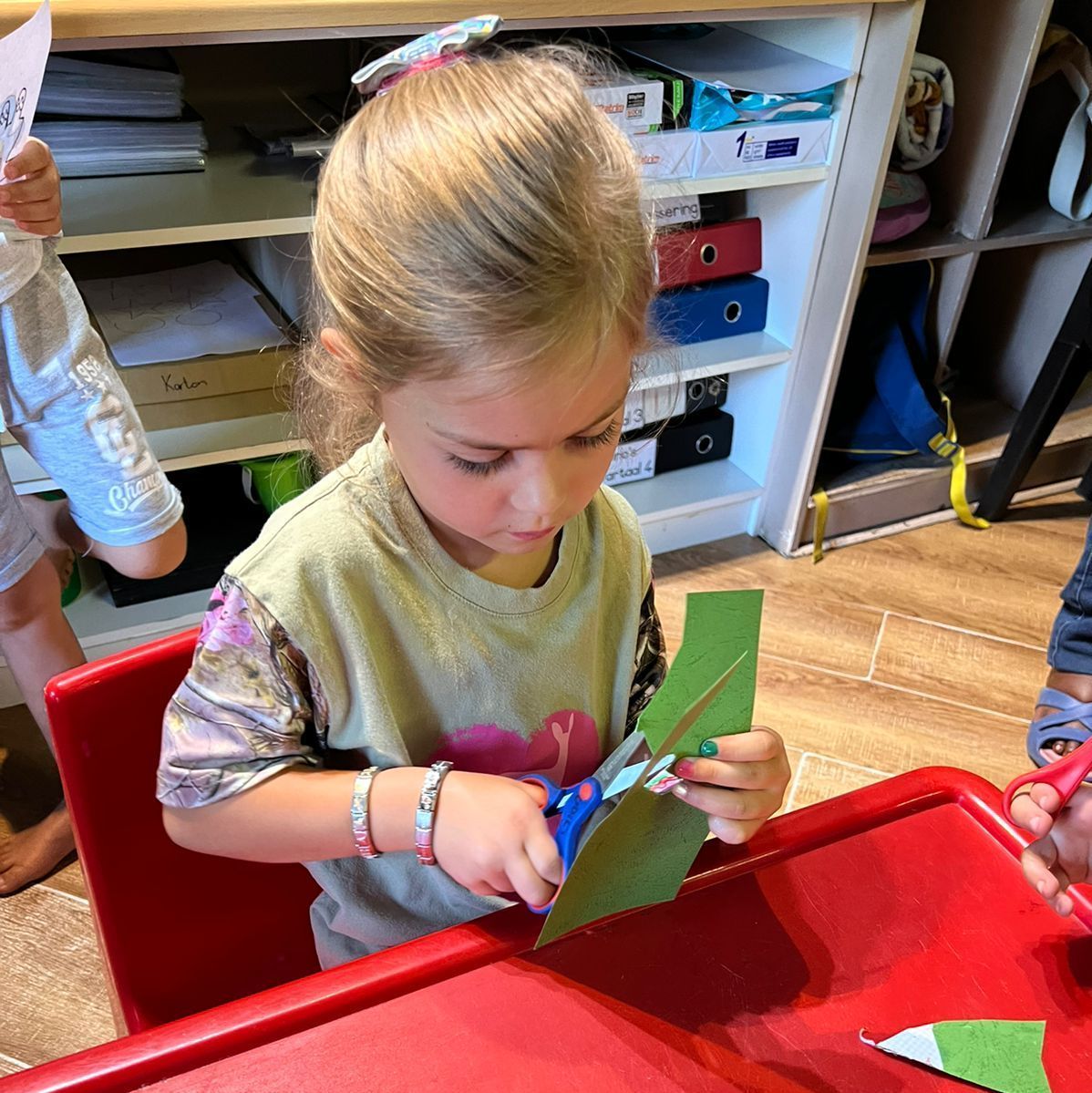 Child sitting at a red table, cutting green paper with scissors. Beige shirt, blonde hair, and a pink bracelet.