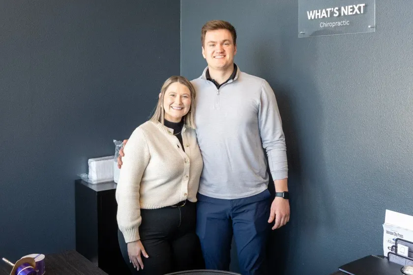 Two people posing indoors in front of a dark wall with a small “WHAT’S NEXT?” sign.