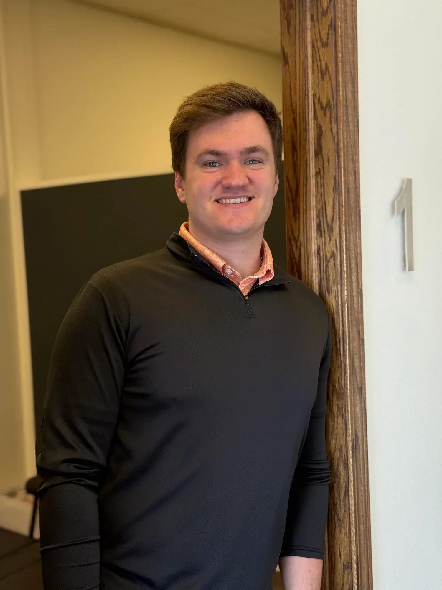 Person in a black shirt standing indoors beside a wooden doorframe, smiling at the camera.