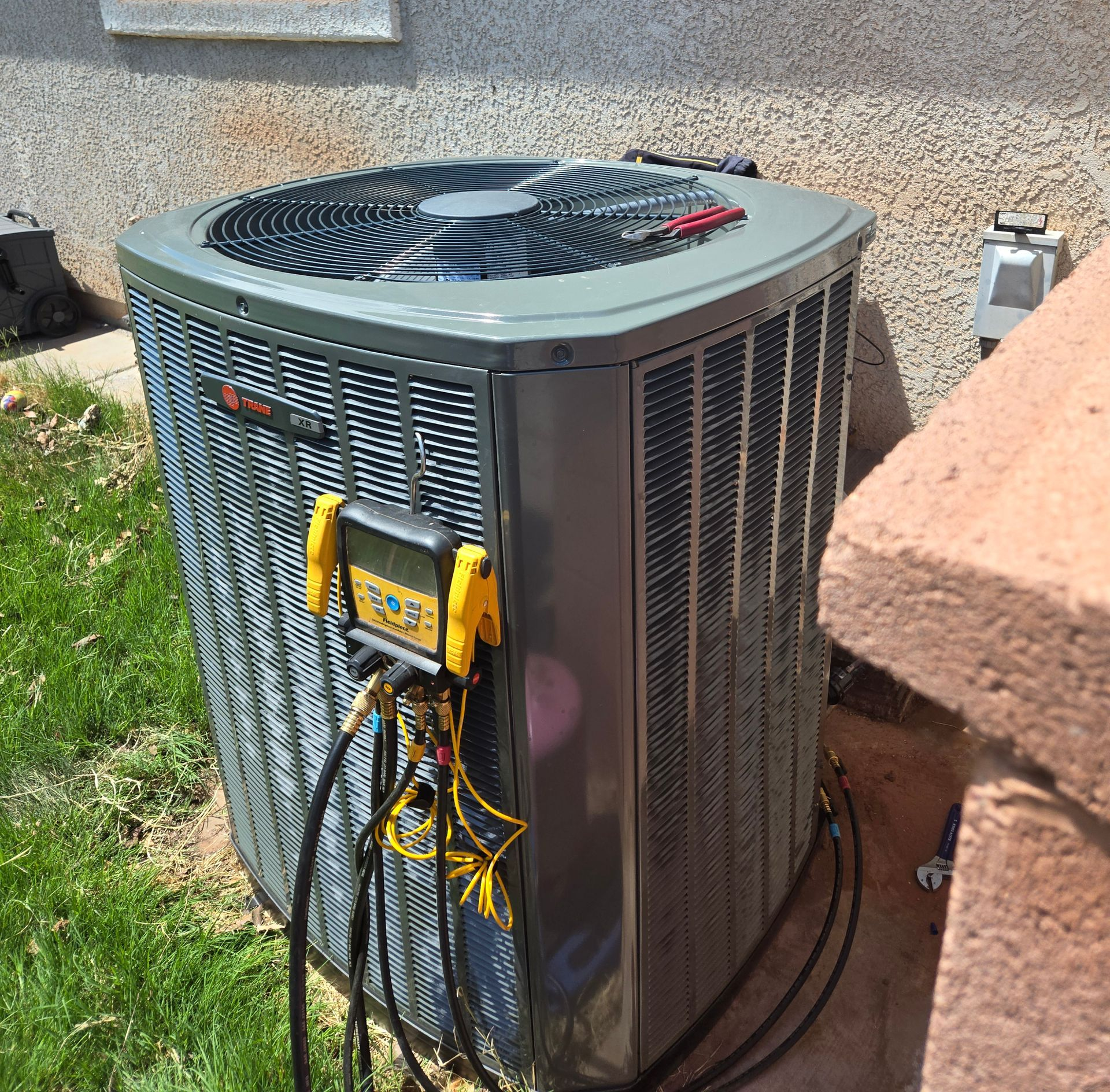 Outdoor HVAC unit with gauges and hoses attached beside a house wall and grass yard
