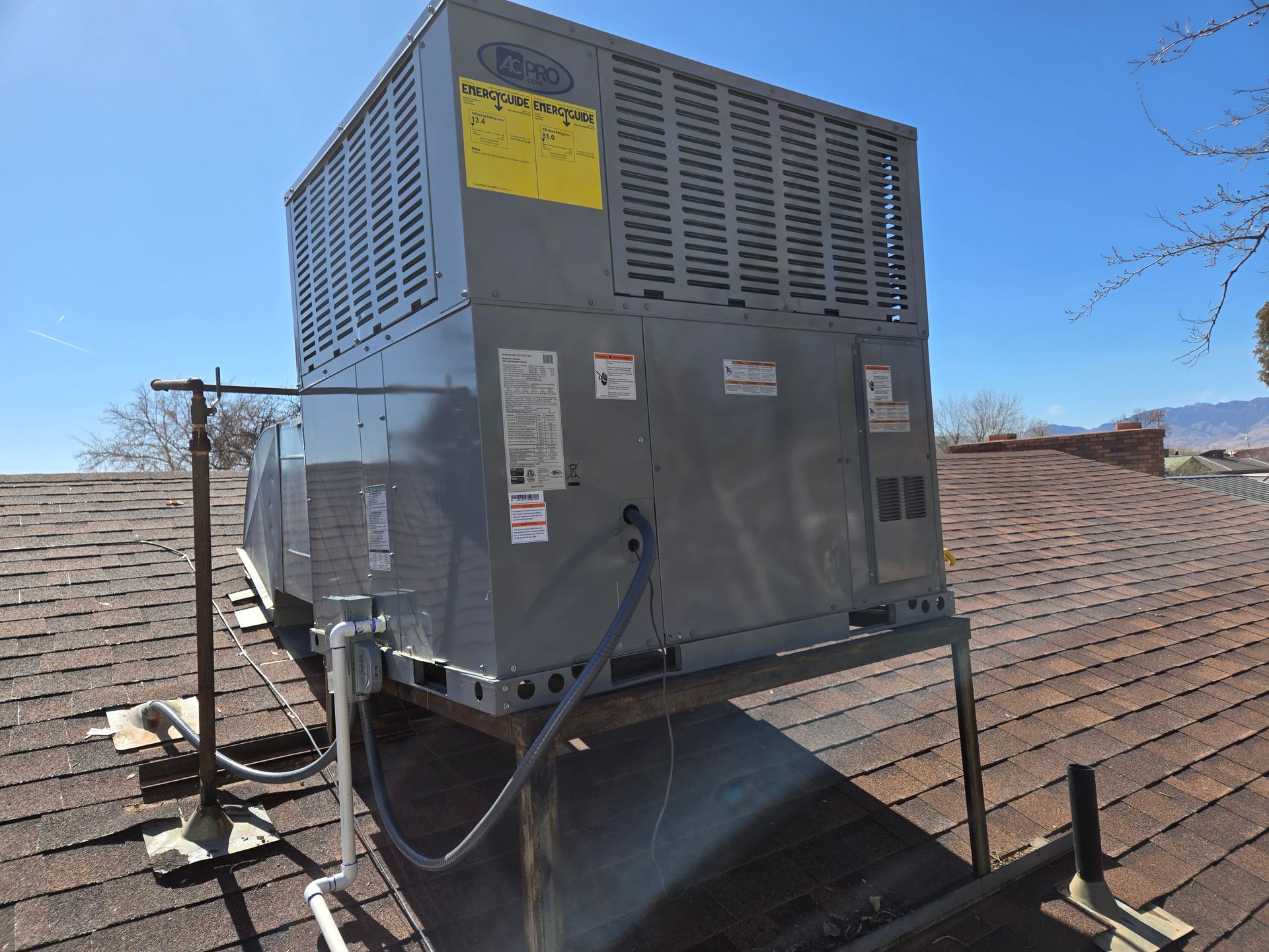 Metal HVAC unit on a rooftop beside vents, with a yellow warning label and mountains in the distance