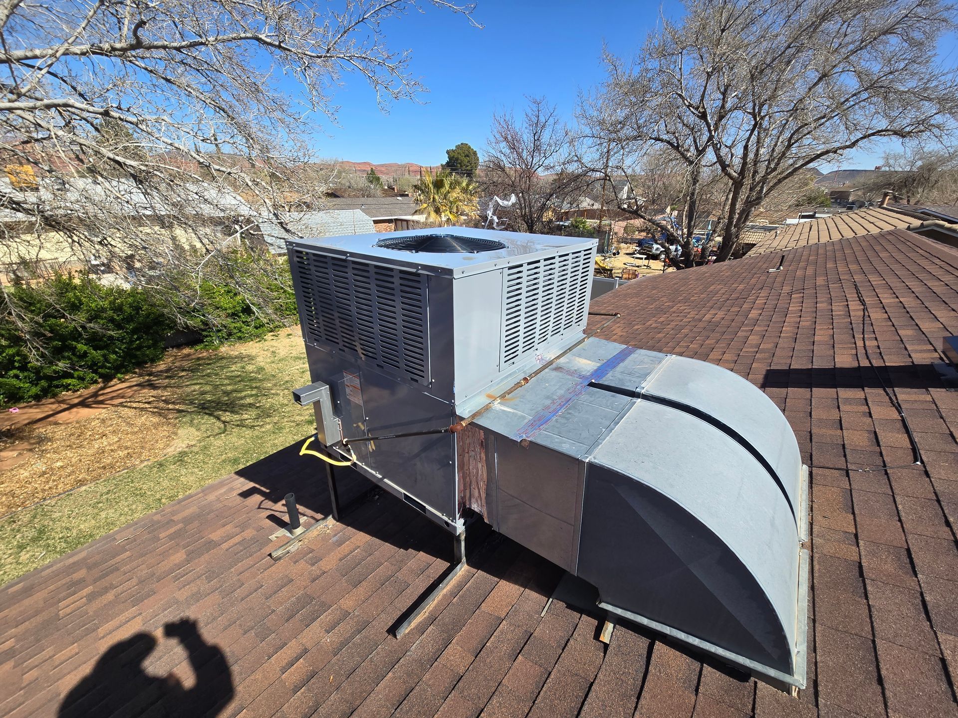 Roof-mounted HVAC unit with curved silver duct on a shingled rooftop under a blue sky
