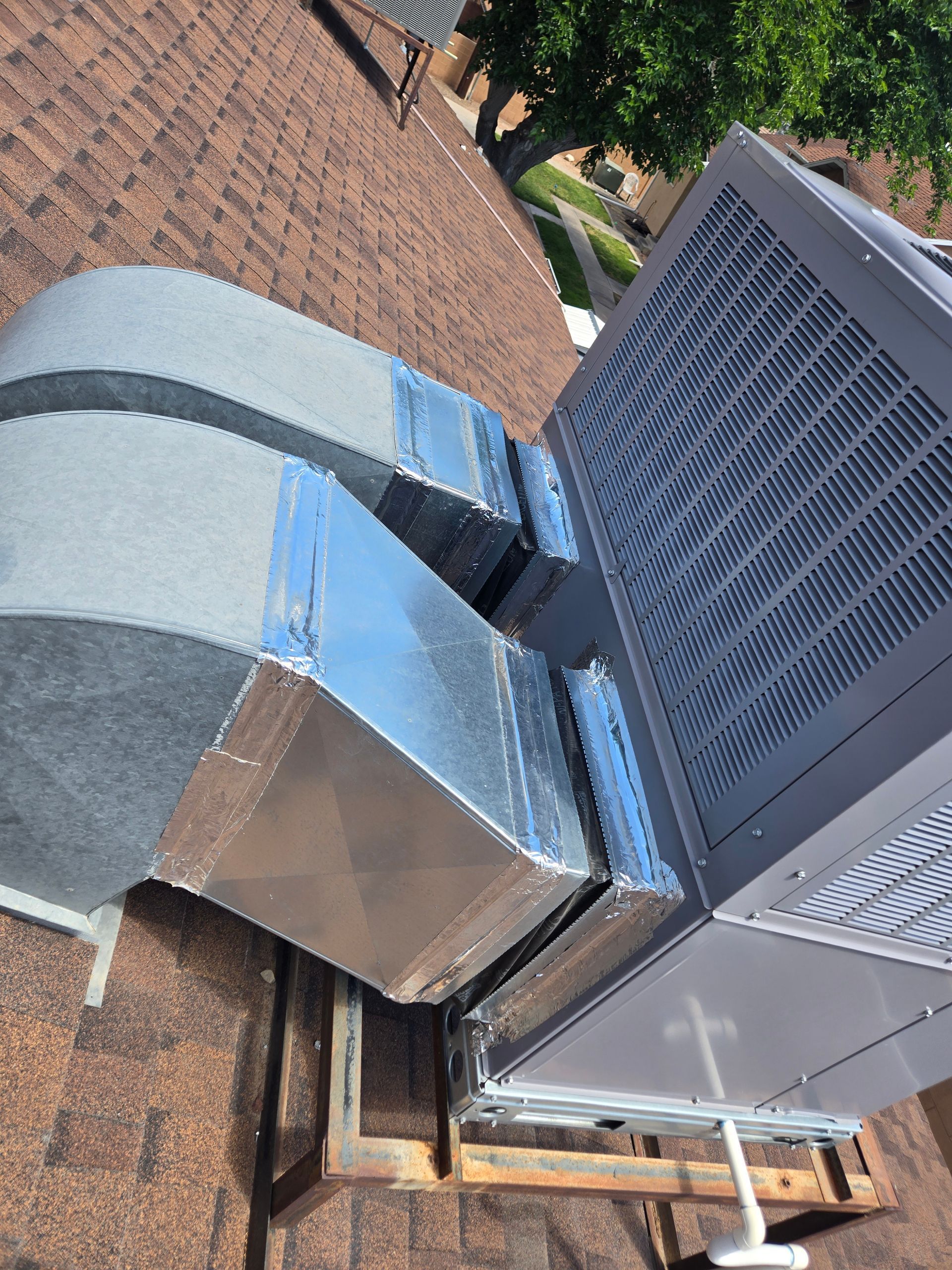 Rooftop HVAC unit with silver ductwork beside a brick building and shingles