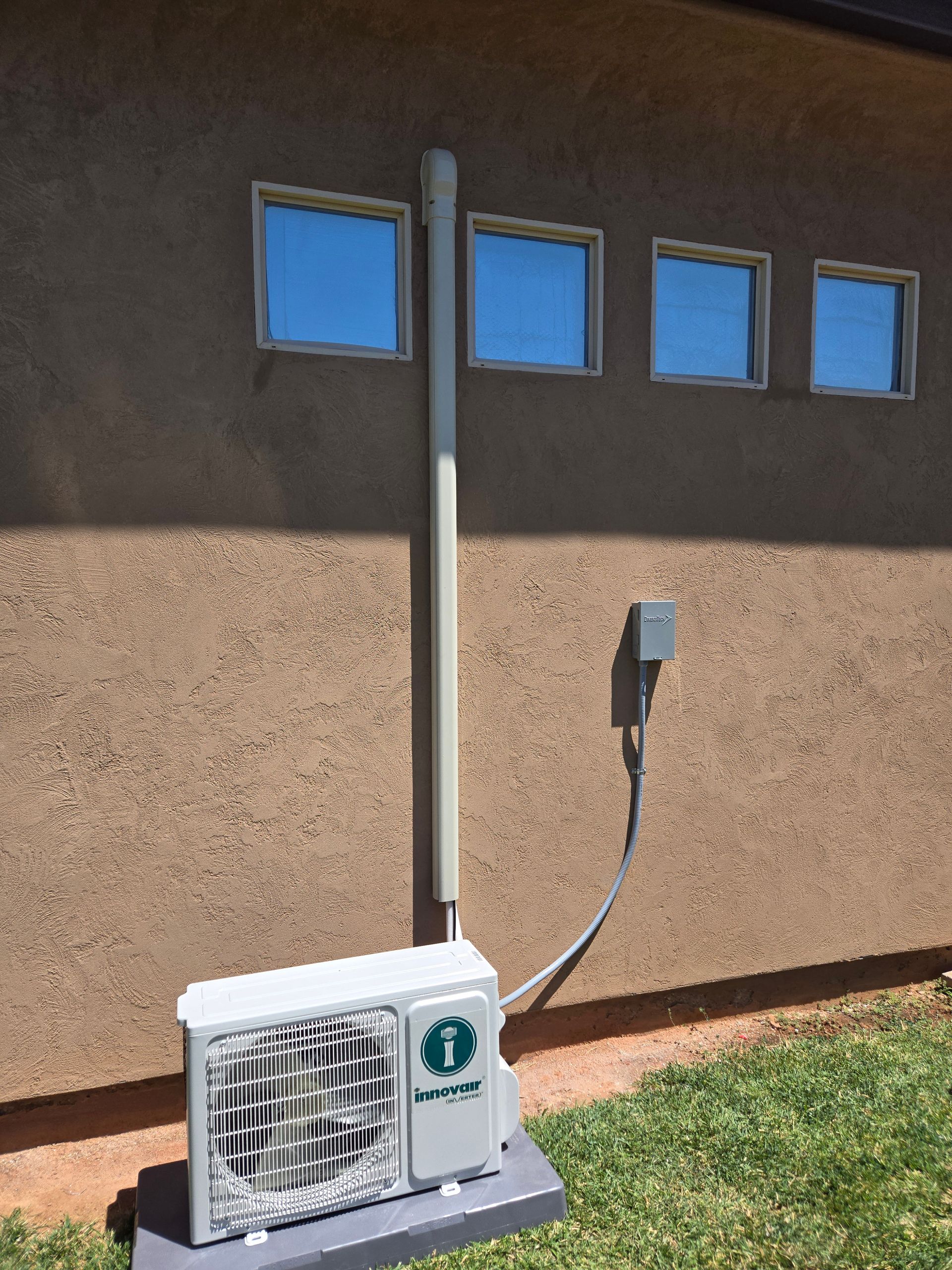 Outdoor HVAC unit beside a building wall with a vertical vent pipe and electrical conduit