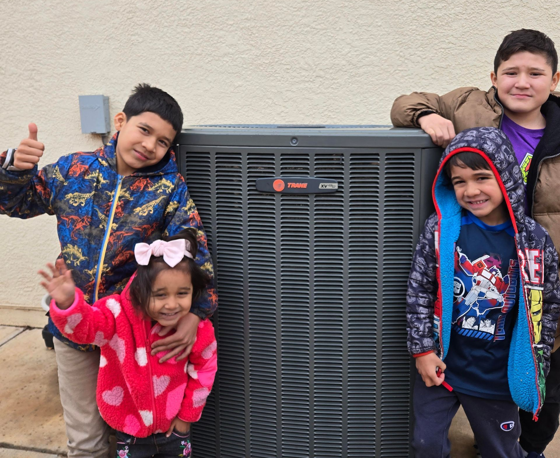 Four children posing and smiling beside a large outdoor HVAC unit against a beige wall.