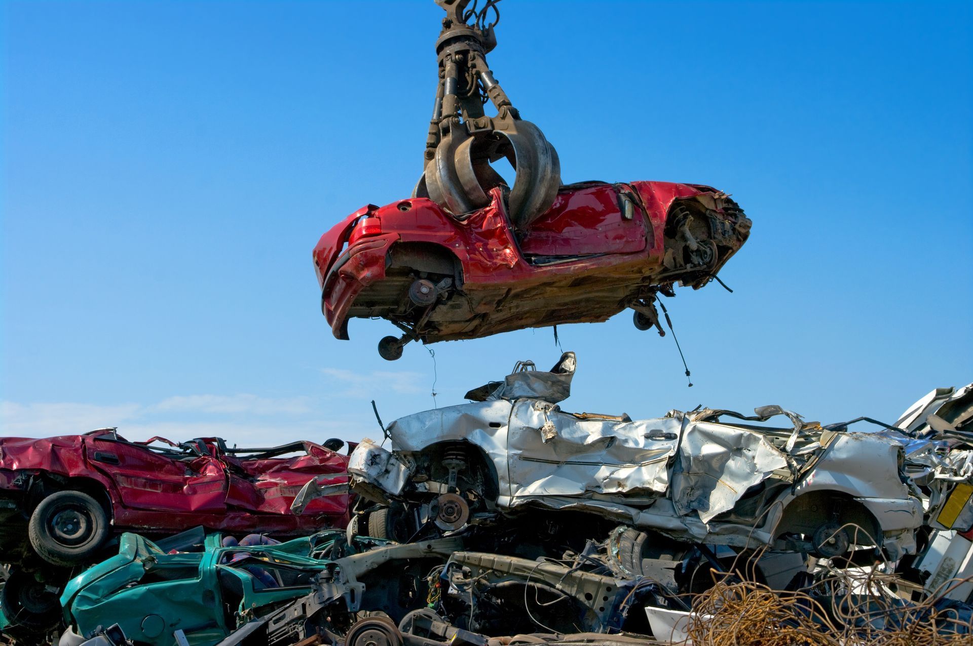 Una gru solleva un'auto rossa schiacciata sopra un cumulo di veicoli distrutti sotto un cielo azzurro.