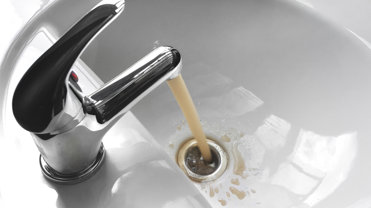 Water streaming from a chrome faucet into a white sink drain, with a slight brown tint.