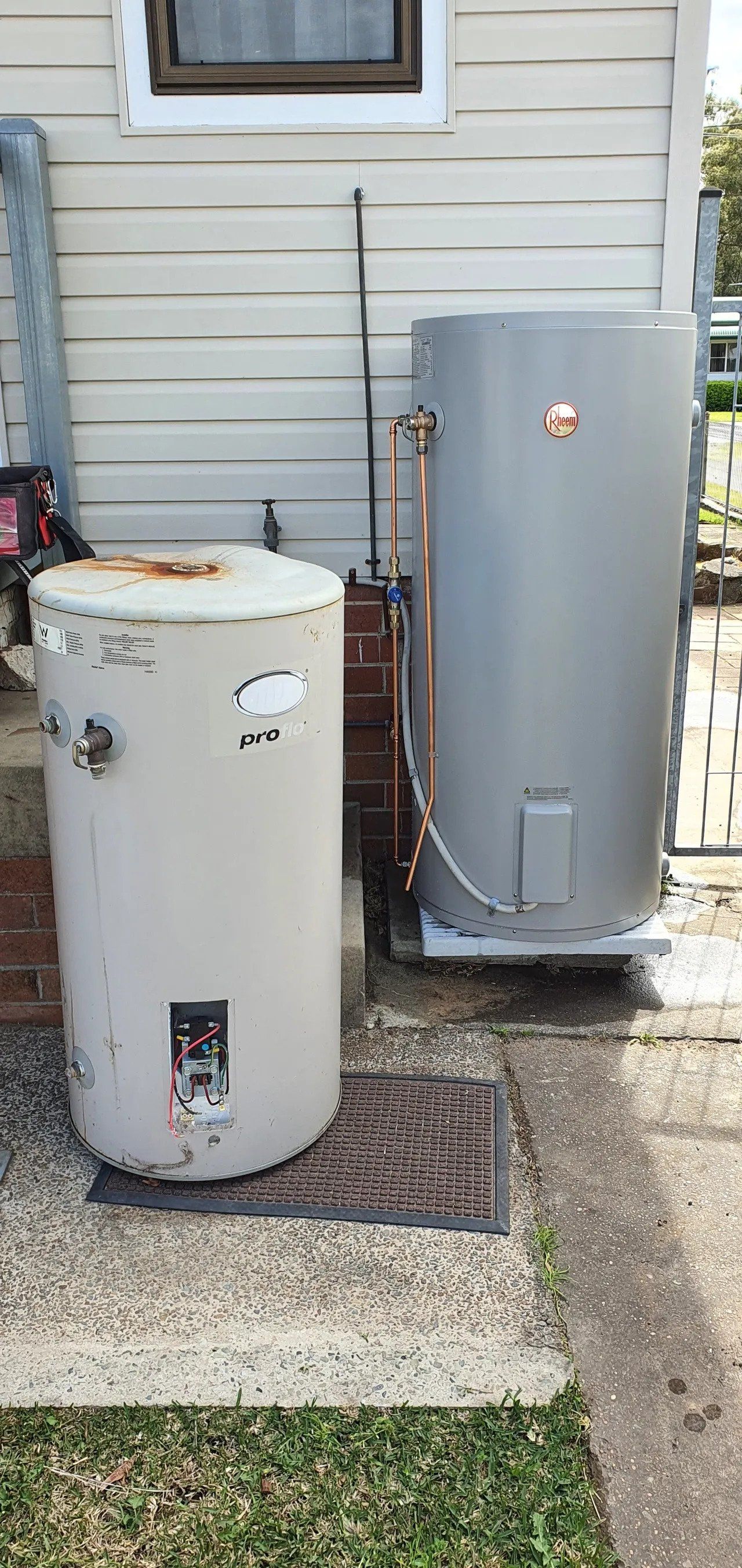 Two outdoor utility tanks beside a house, one white and one gray, on a gravel pad.