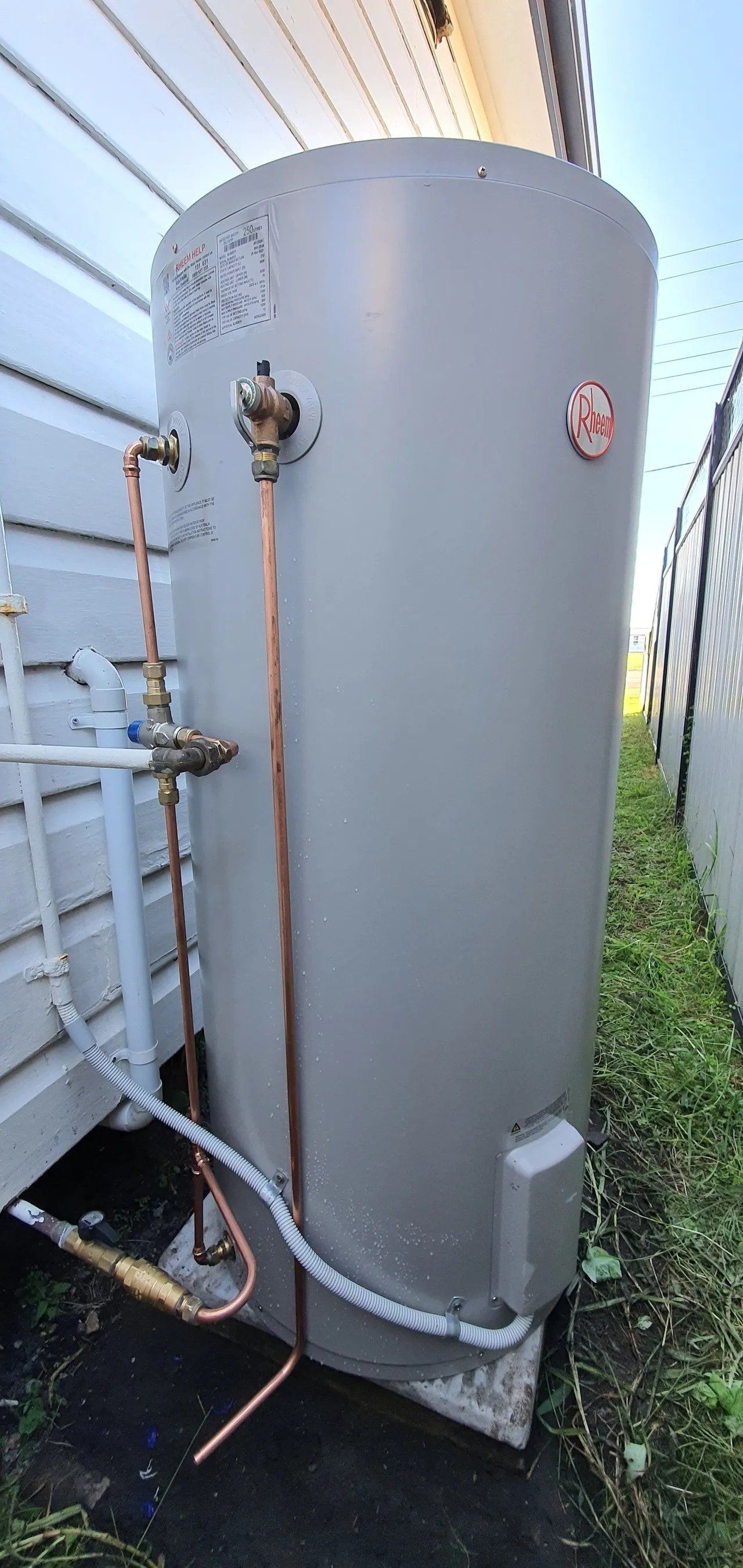 Outdoor gray cylindrical water heater beside a house, with exposed pipes and grass nearby