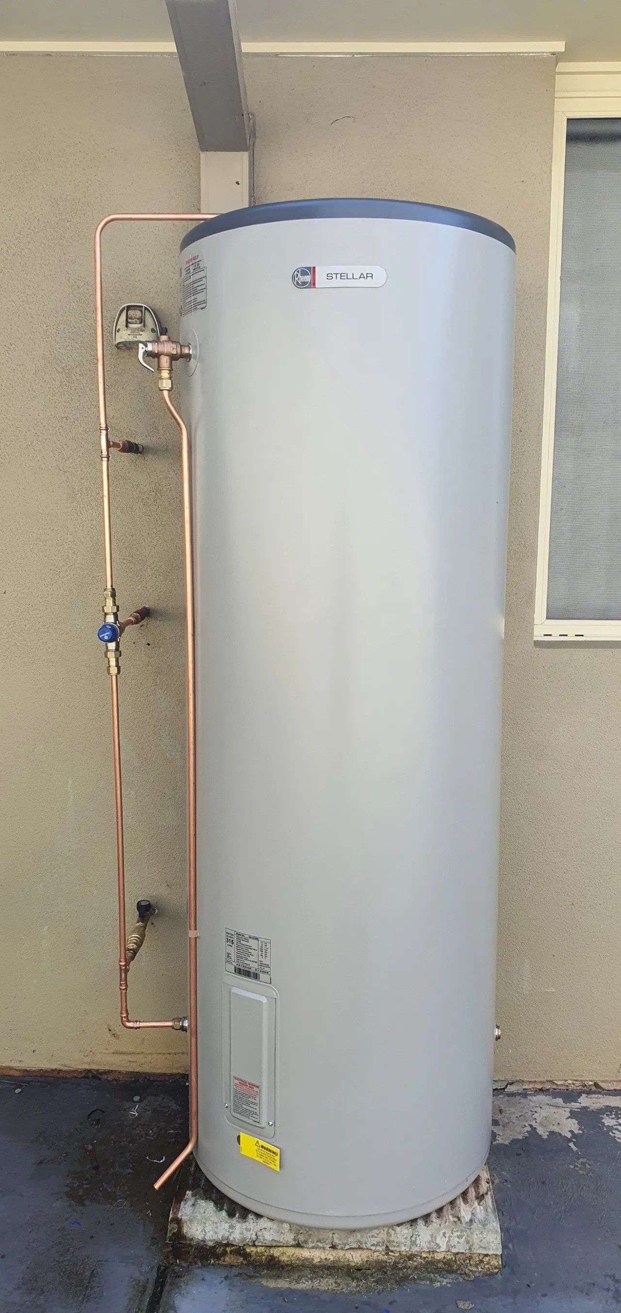 Tall white water heater tank in a utility room, with exposed copper pipes and a gray textured wall.