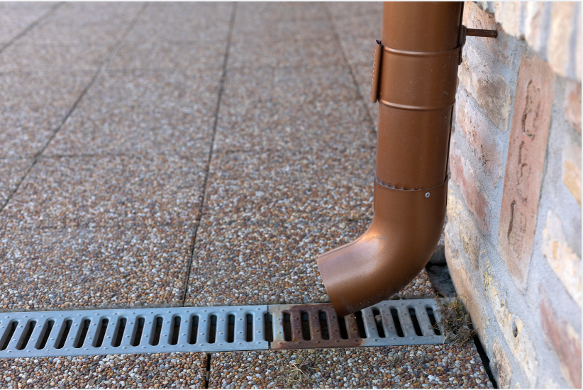 Brown downspout drains rainwater into a metal street grate beside a brick wall.