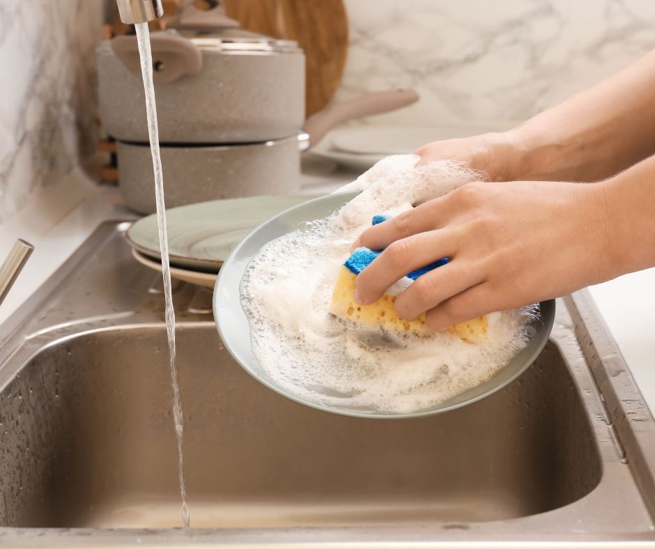 Hands washing a dish in a kitchen sink with running water and soap suds