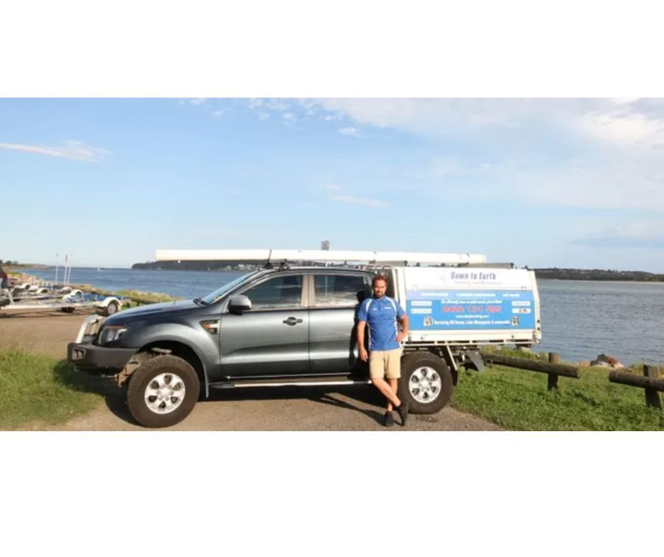 Woman standing by a black pickup truck with a blue sign, parked near the water on a sunny day