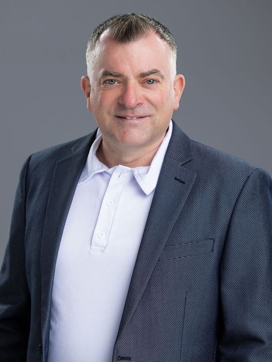 Man in navy blazer and white polo smiling, against a gray background.