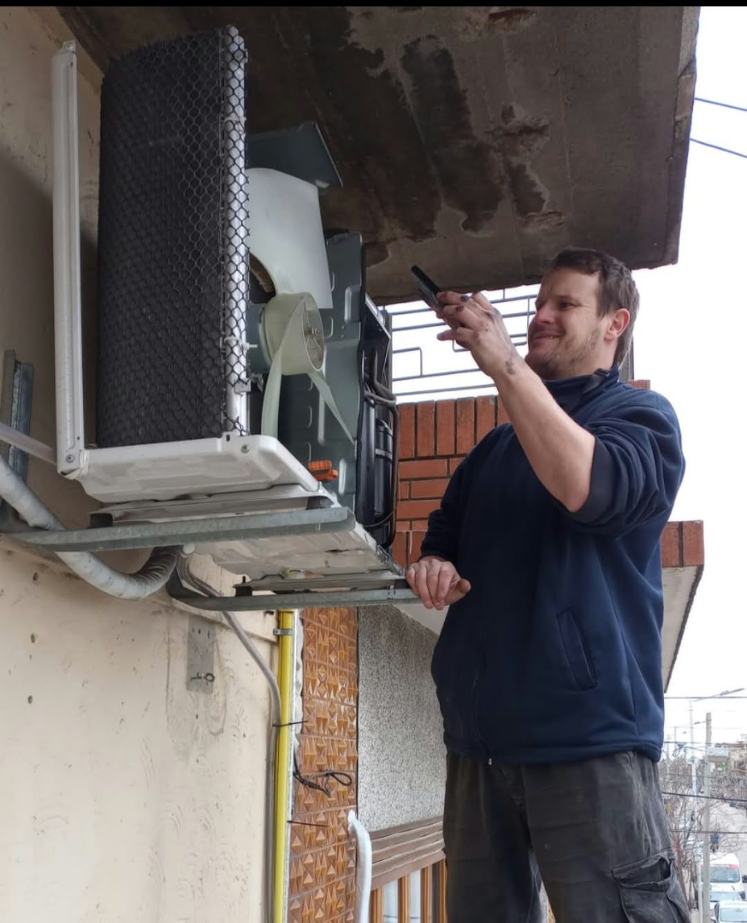 Hombre ajustando una unidad de aire acondicionado exterior en la pared de un edificio, debajo de un balcón