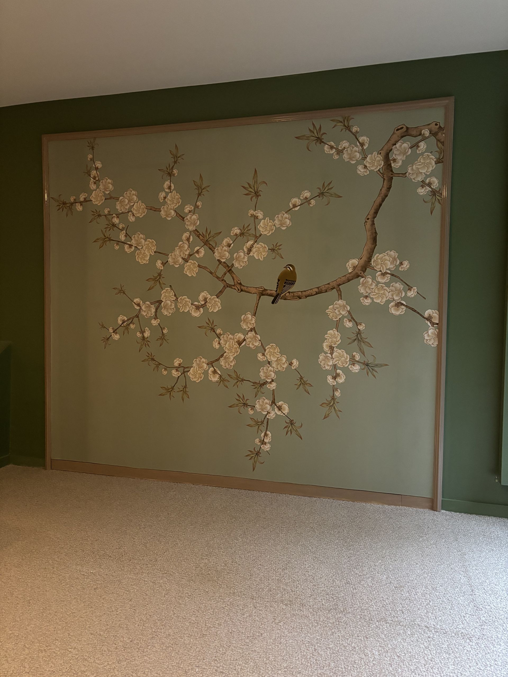 Grande fresque murale représentant une branche d'arbre en fleurs sur un mur vert pâle, au-dessus d'une moquette beige.