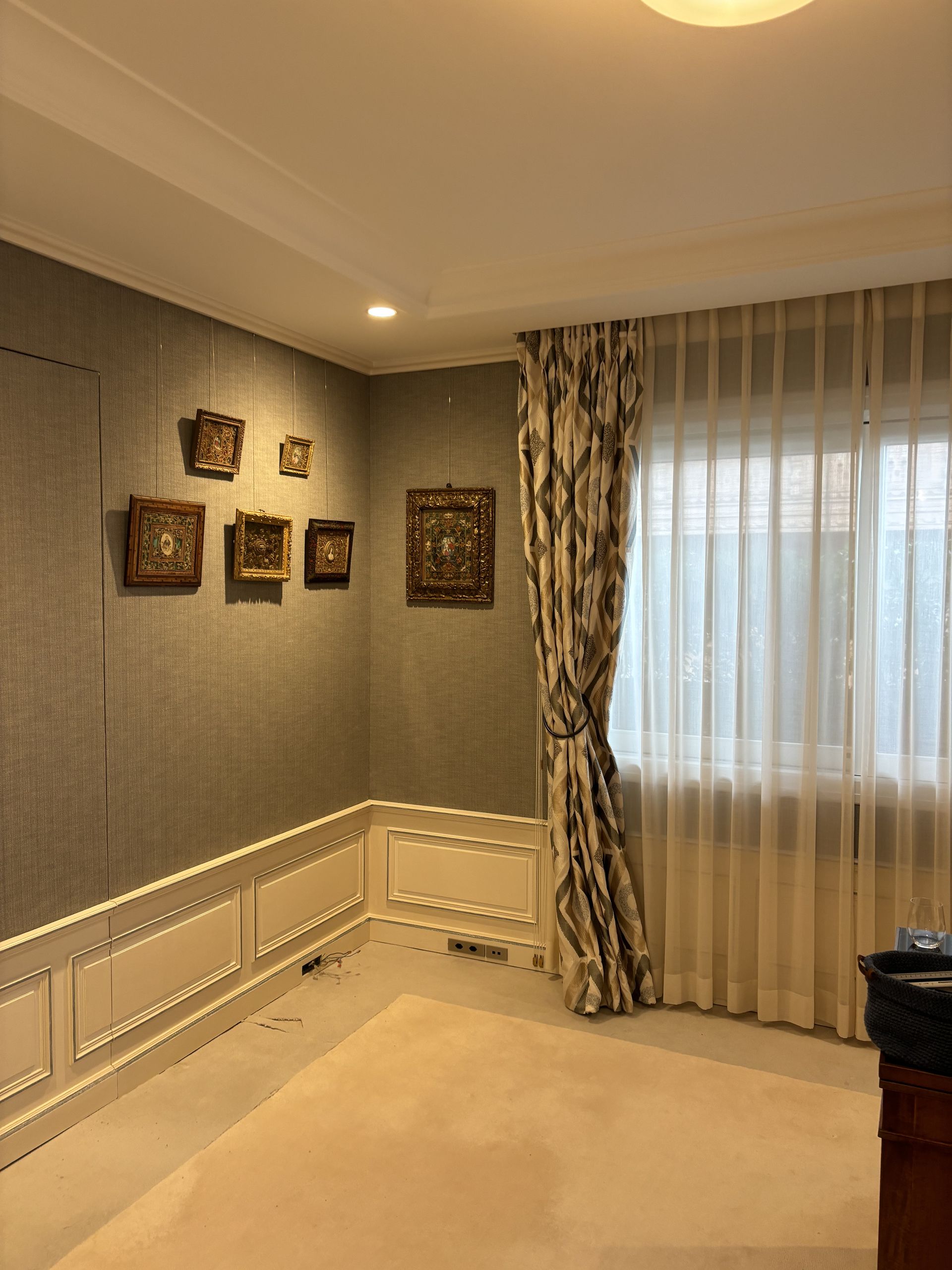 Pièce beige vide avec des tableaux encadrés, du papier peint à motifs et des voilages à la fenêtre.