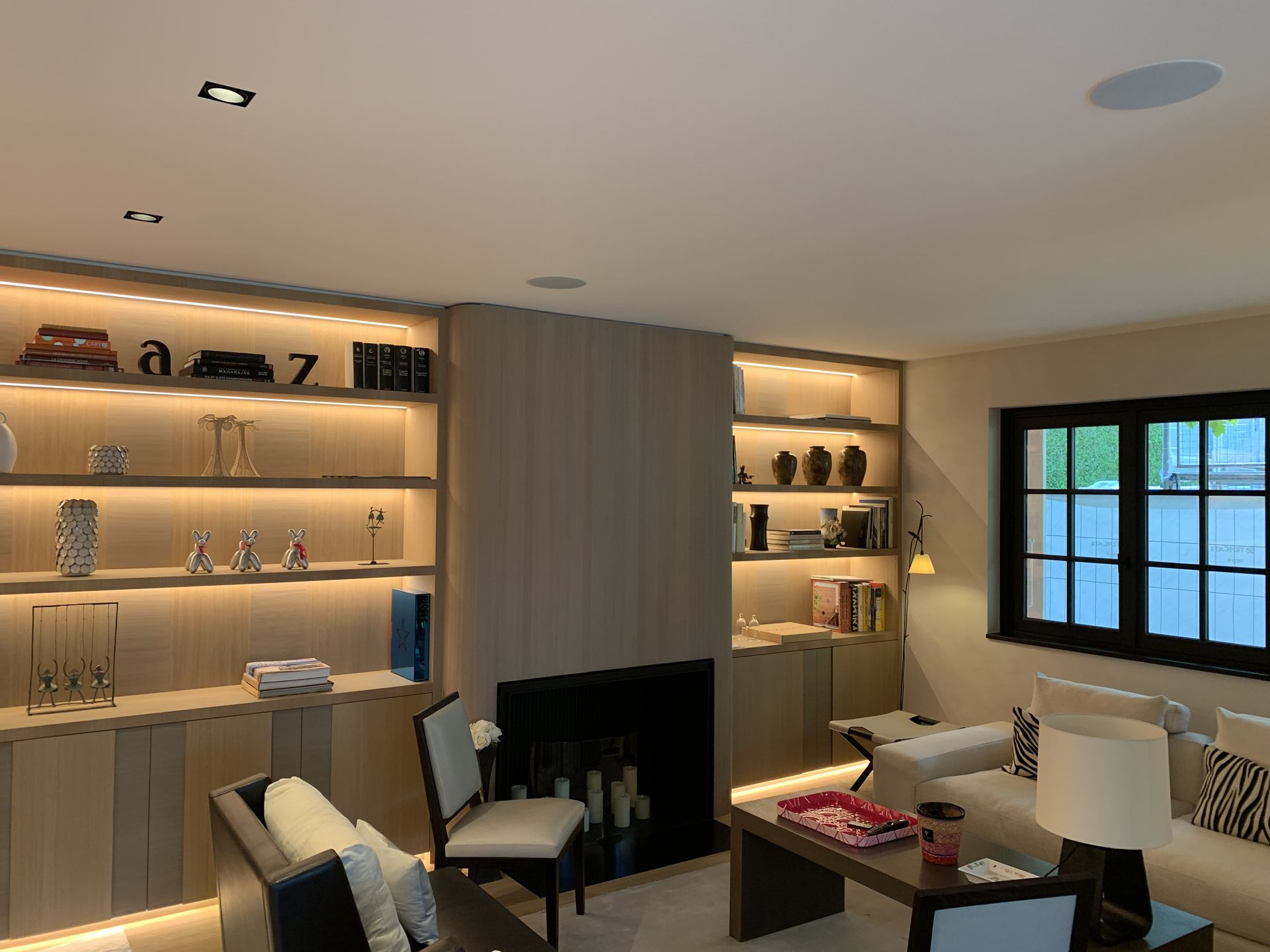 Salon moderne avec étagères éclairées intégrées, cheminée, fauteuils et fenêtre à cadre noir.