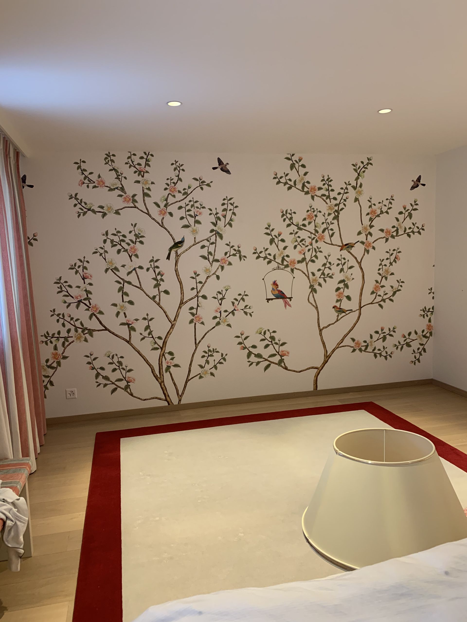 Chambre calme avec une fresque murale florale, un tapis crème et des rideaux à rayures ; un abat-jour est posé au sol.