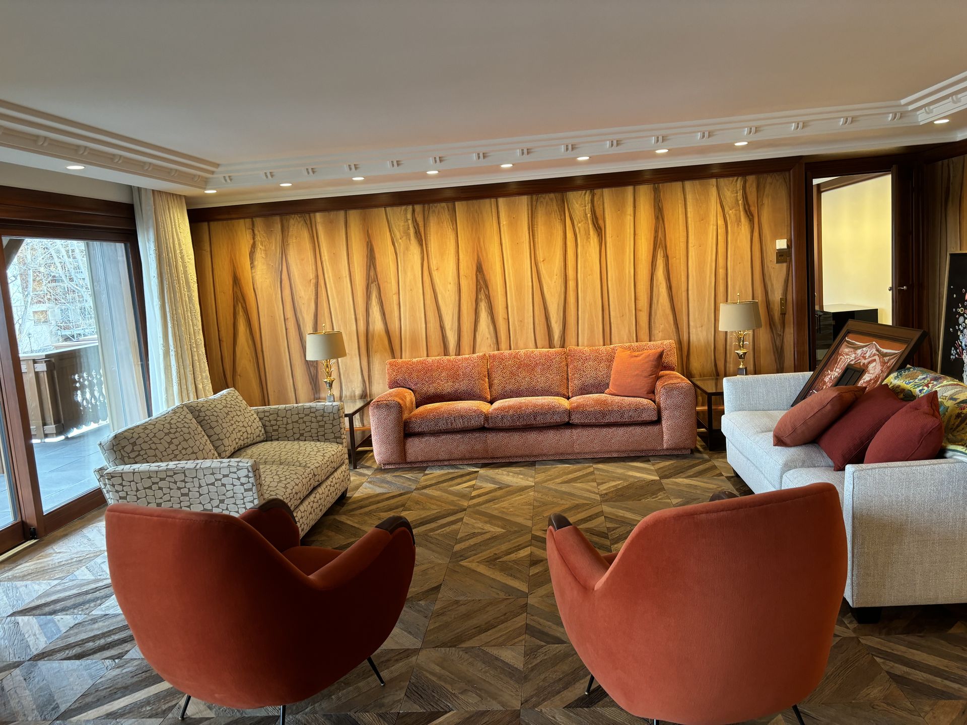 Salon moderne avec canapés orange et blancs, fauteuils rouges, mur lambrissé et grandes fenêtres