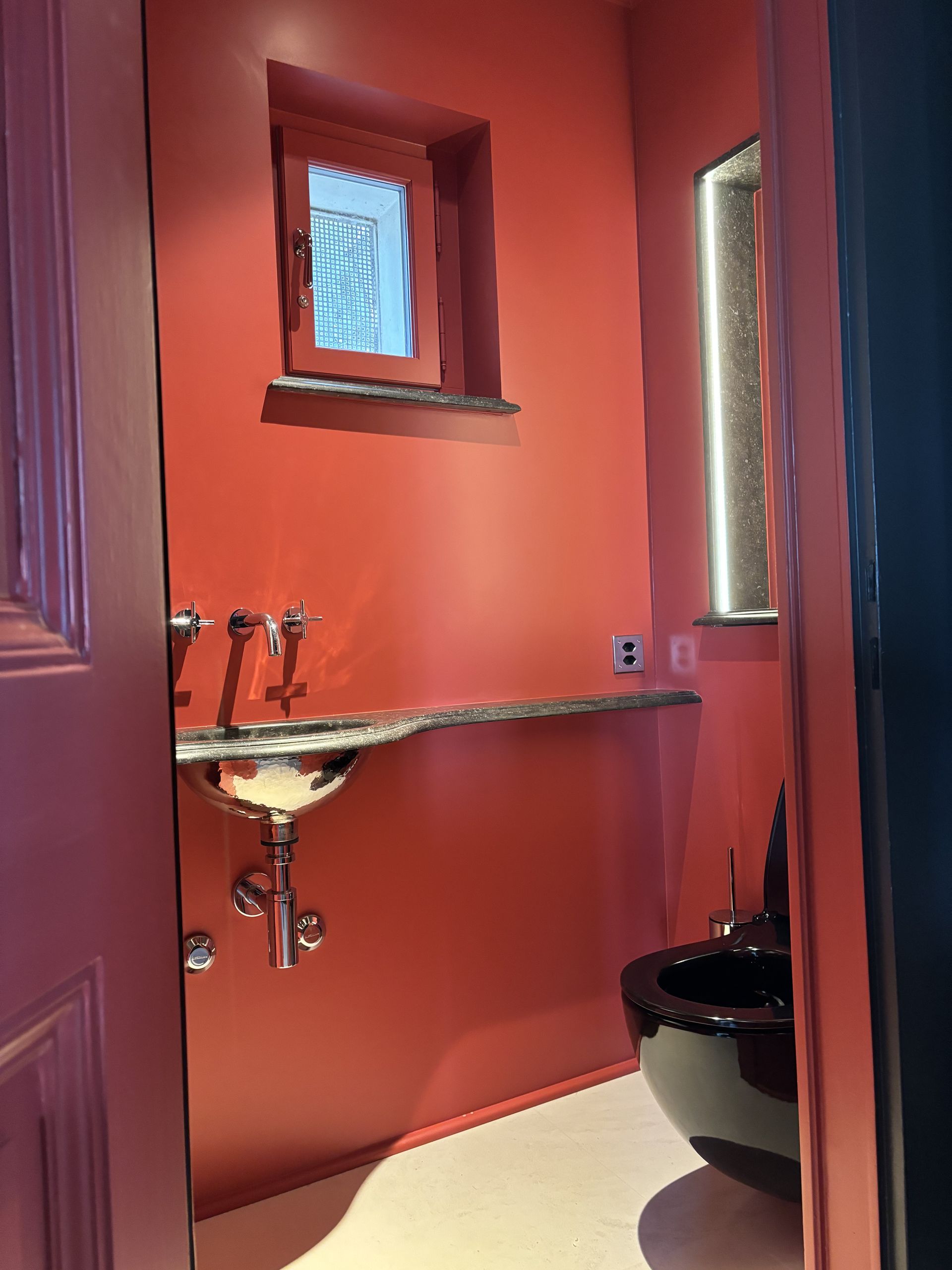 Petite salle de bains rouge avec lavabo mural, fenêtre étroite et partie des toilettes visible.
