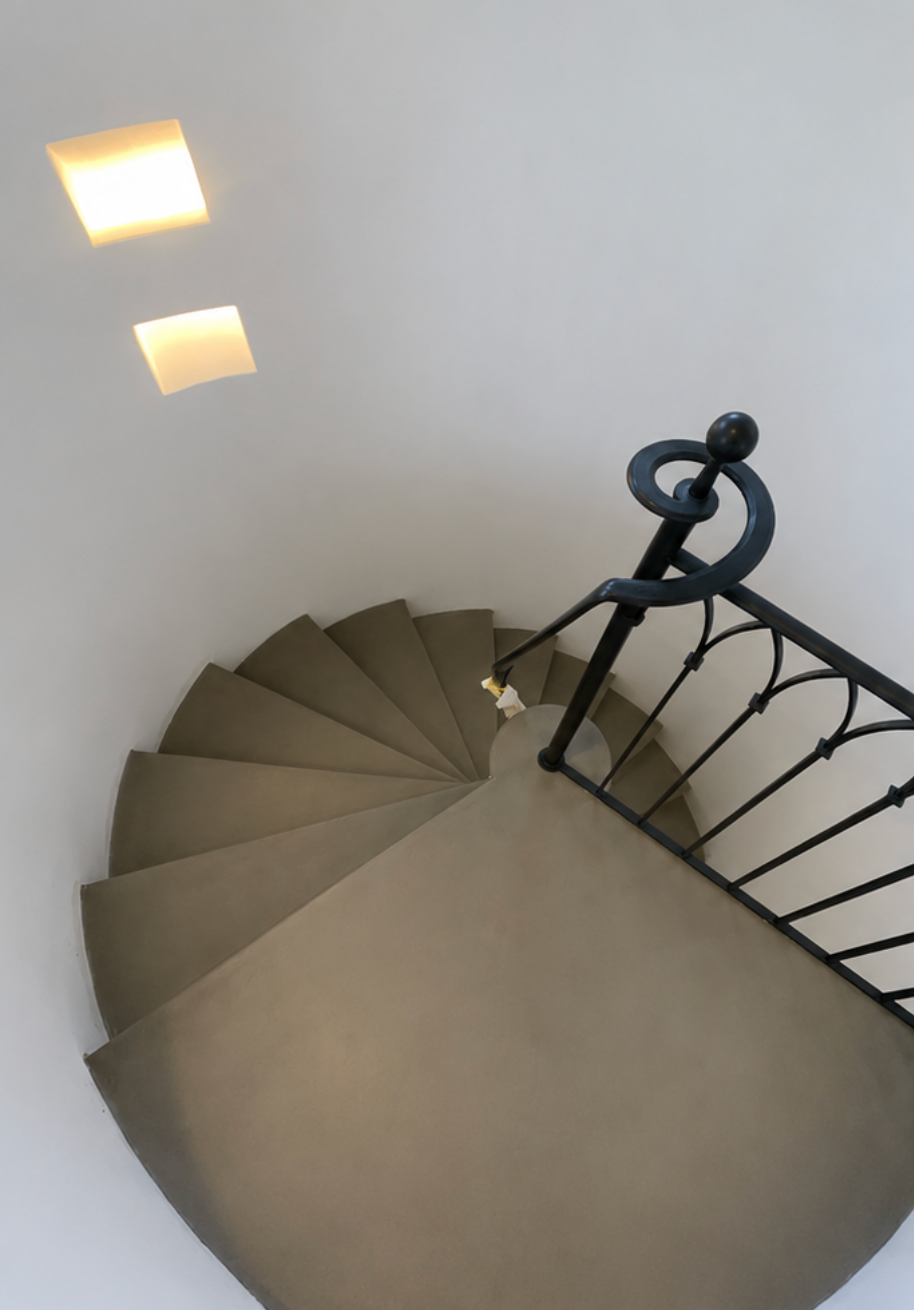 Escalier en colimaçon avec marches beiges, rampe noire et deux plafonniers dans un intérieur blanc.