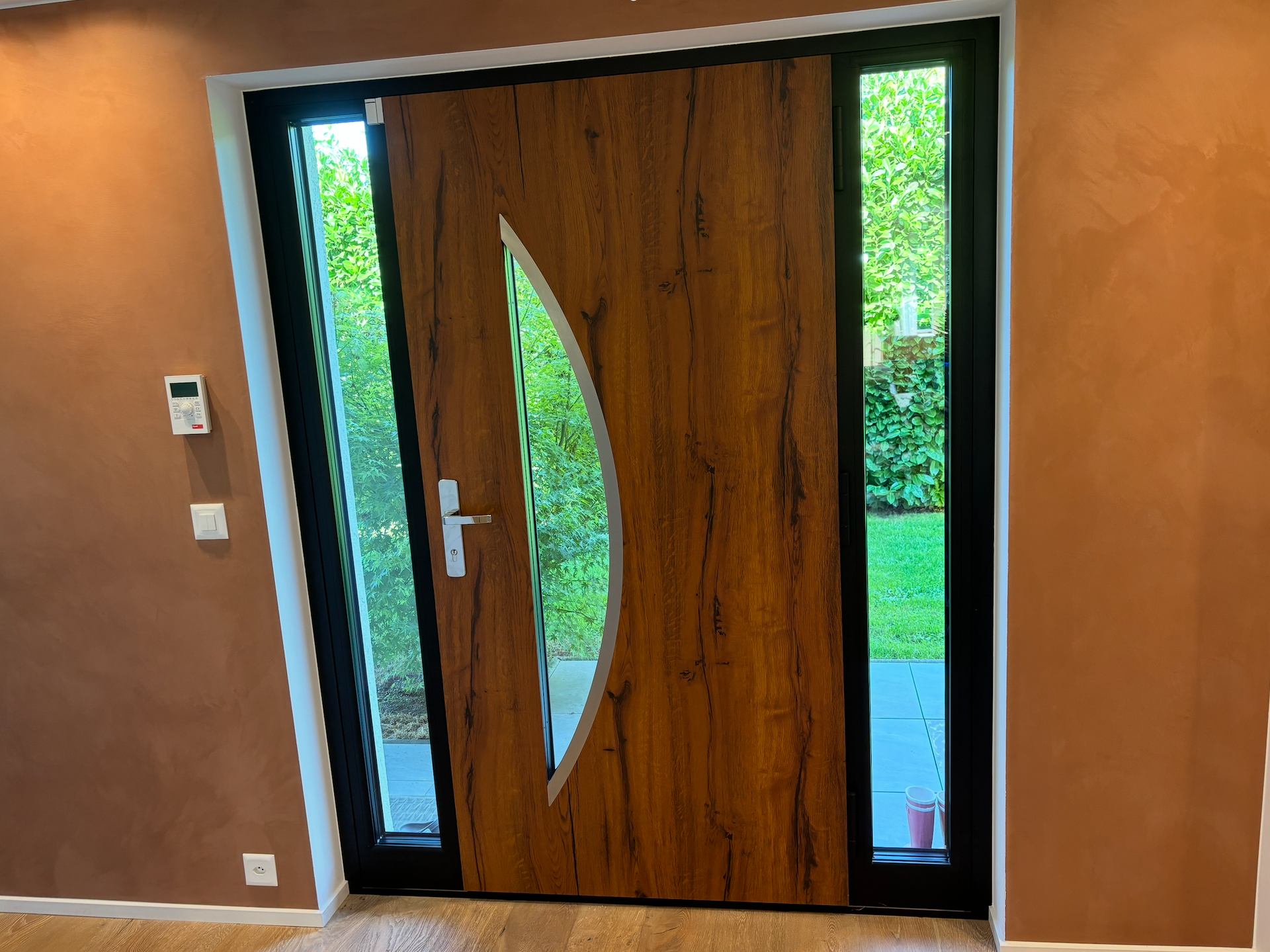 Porte d'entrée en bois avec un panneau de verre incurvé, flanquée d'étroites fenêtres dans un hall d'entrée beige.