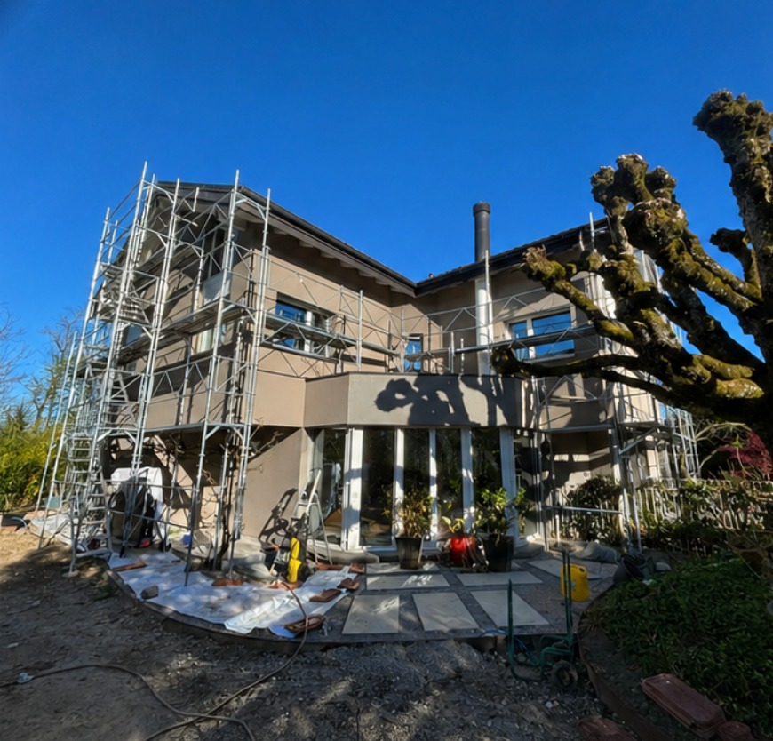 Extérieur d'une maison avec échafaudages, véranda, ciel bleu et arbre taillé devant.