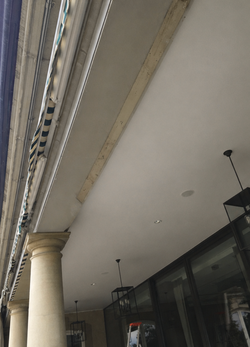 Passage couvert avec une colonne en pierre, des vitrines et des suspensions noires sur un plafond gris.
