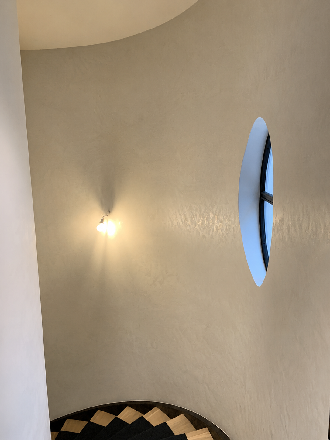 Mur d'escalier blanc incurvé avec une applique murale, fenêtre ovale étroite et sol à damier noir et blanc en dessous