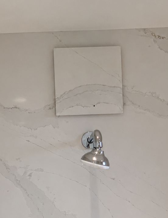 Mur de salle de bain avec un petit panneau carré et une pomme de douche chromée en dessous ; le mur présente des taches d’eau et des fissures.