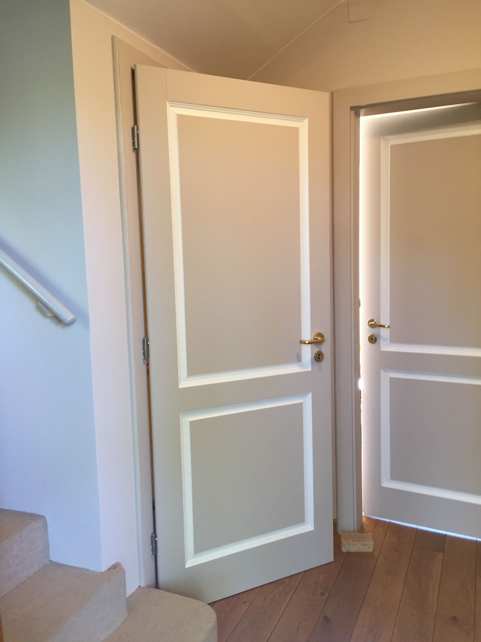 Porte blanche ouverte dans un couloir beige avec parquet et rampe d'escalier sur la gauche
