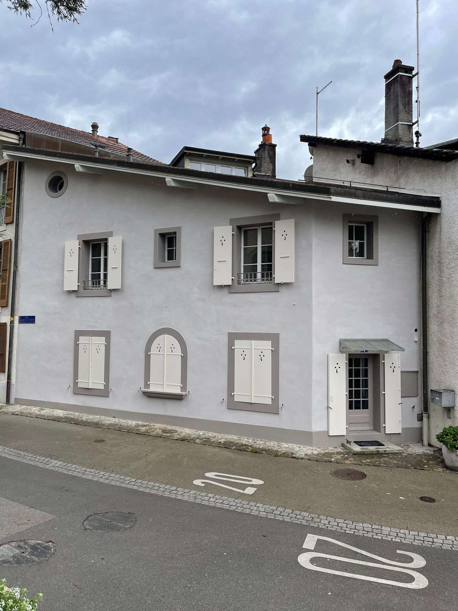 Maison grise à deux étages avec des volets blancs, située au bord d'une rue en pente sous un ciel nuageux.