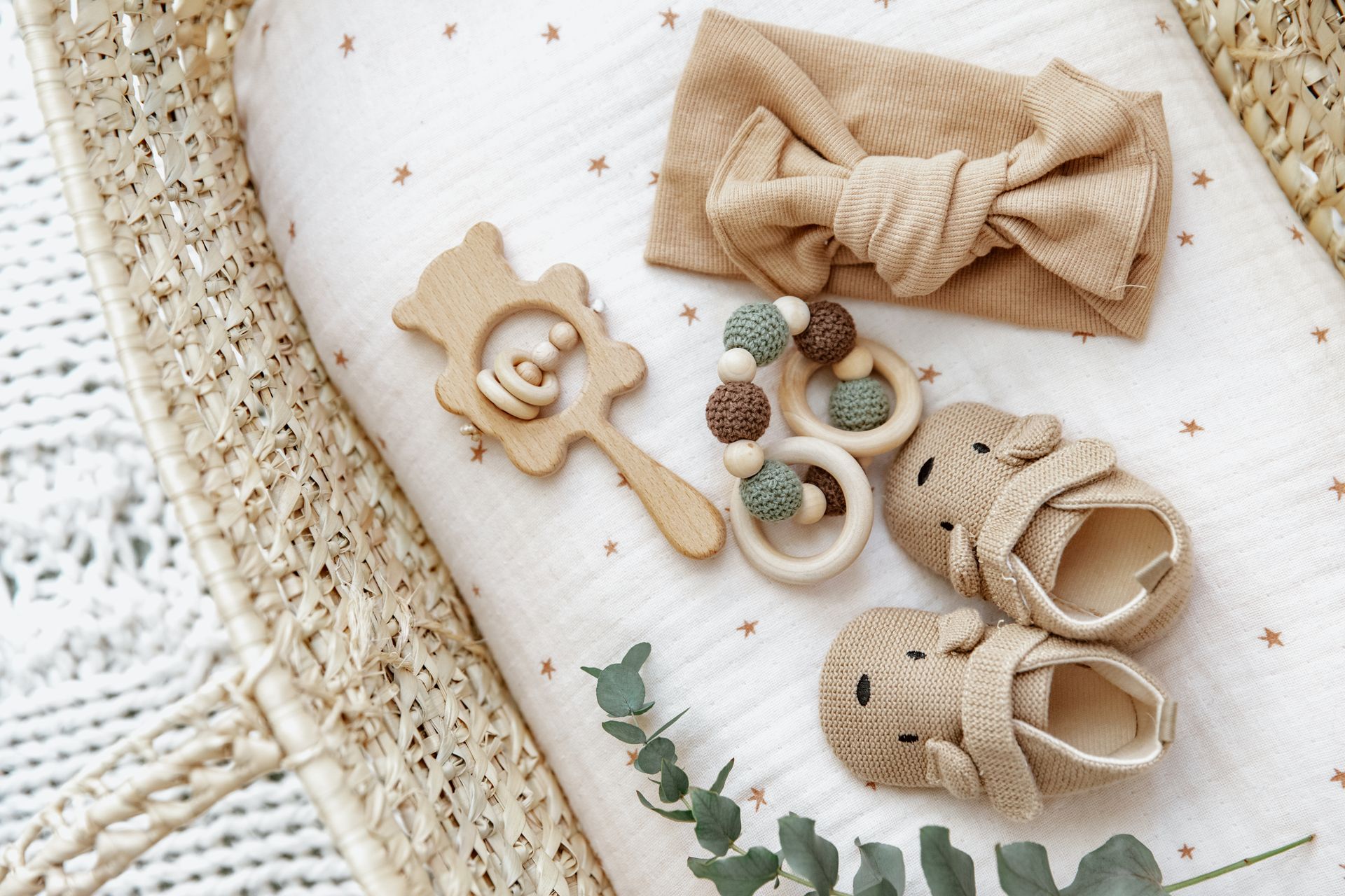 Baby accessories arranged on a soft white blanket: bow headband, wooden toys, teethers, and knit booties Baby accessories on a white blanket: beige bow headband, wooden rattle, pacifier clips, and tiny sandals.