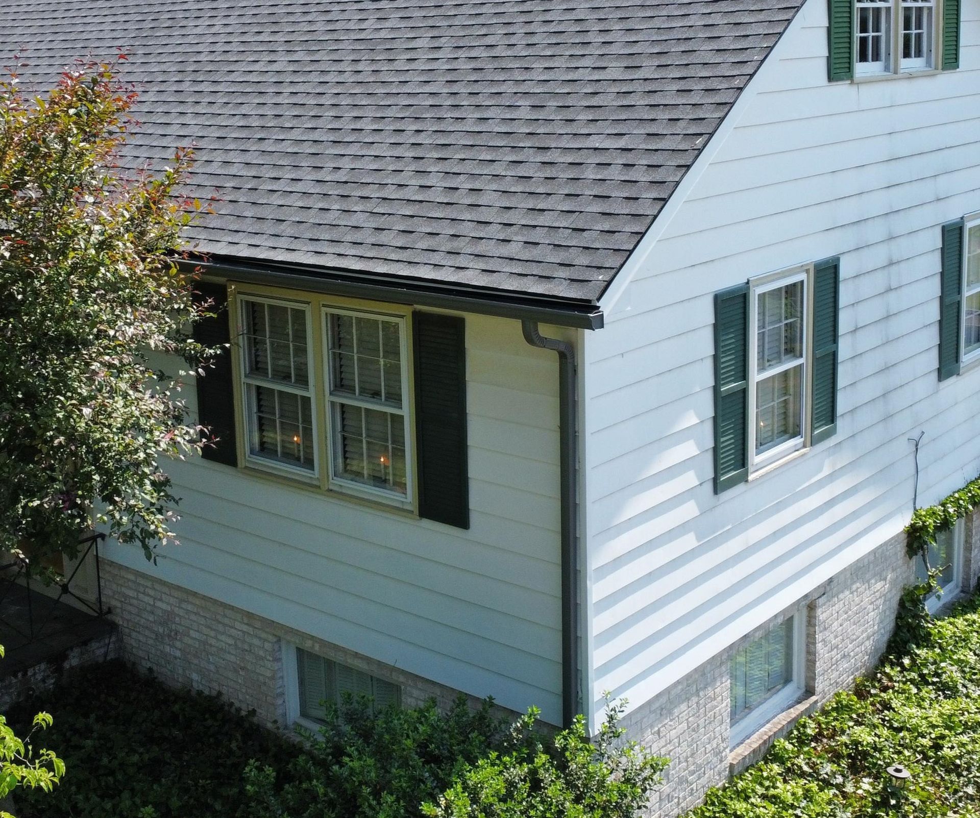 White house exterior with gray shingled roof, green shutters, and a small tree in the yard