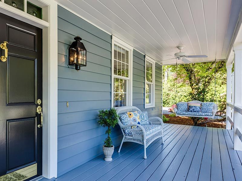Covered porch with black door, blue siding, white chair, and cushioned seating in sunlight