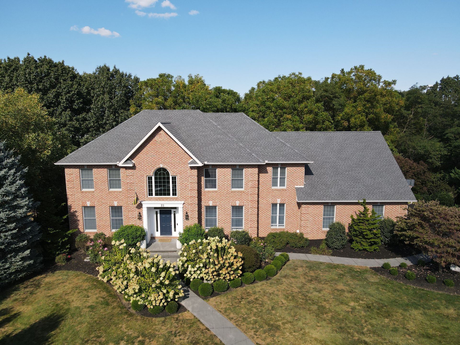 Large brick house with gray roof, white-trimmed windows, and a landscaped front yard under a blue sky