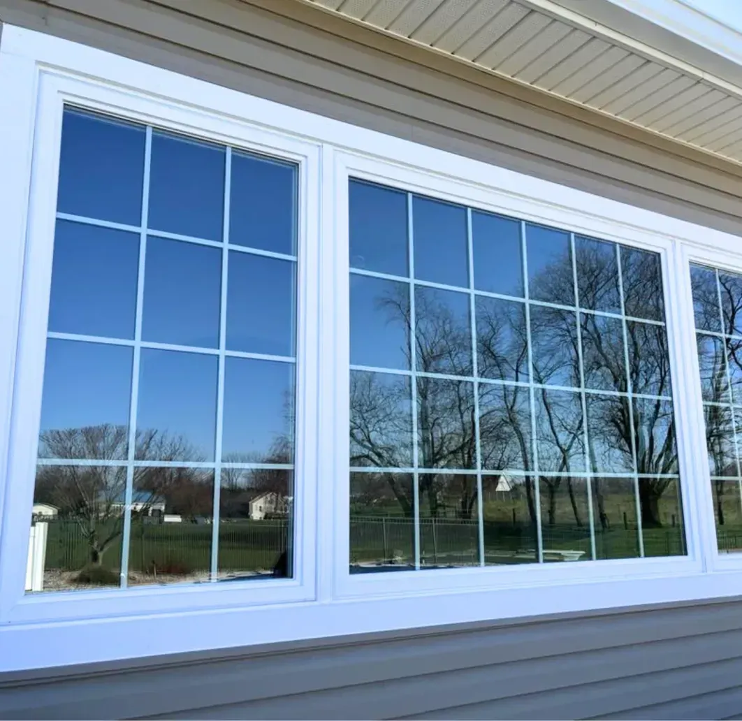 White-framed windows reflecting trees and a blue sky on a beige house exterior