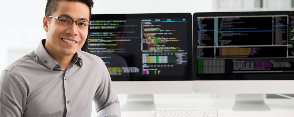 A man is sitting in front of two computer monitors and smiling.