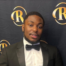 Man in tuxedo with bow tie smiling, in front of a backdrop with a gold