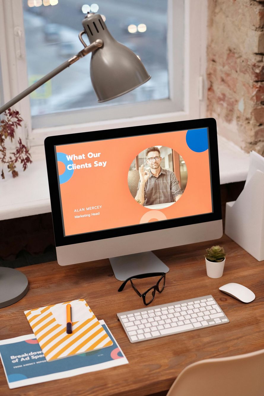 Computer screen displaying presentation, desk with keyboard, glasses, and documents, by window.
