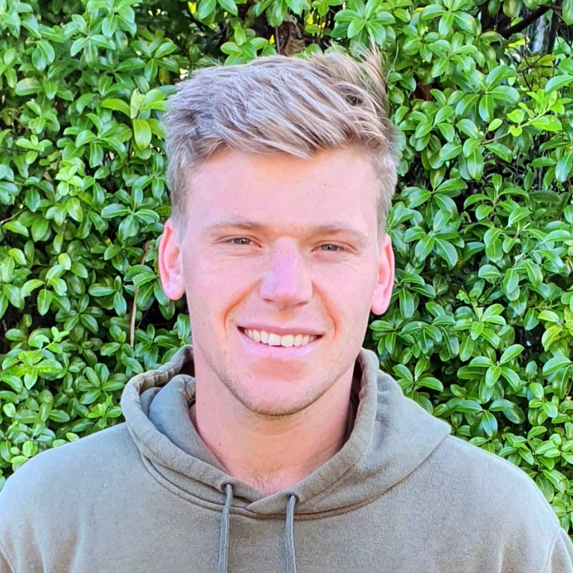 A young man in a hoodie is smiling in front of a bush.