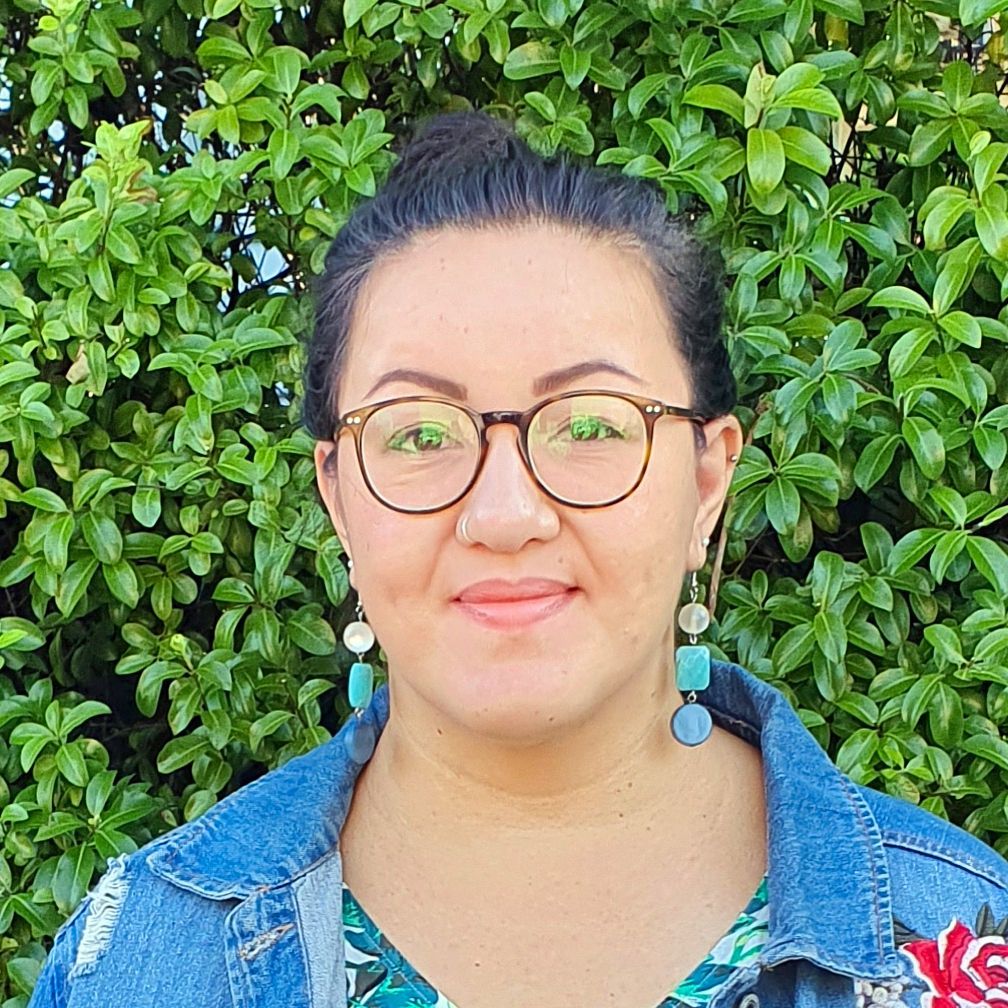 A woman wearing glasses and earrings is standing in front of a bush.