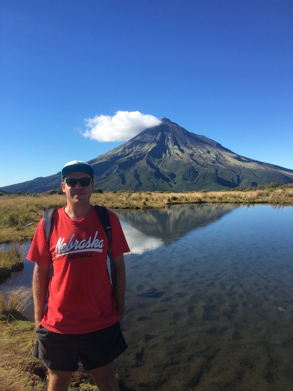 A man in a red shirt is standing in front of a mountain and a lake.