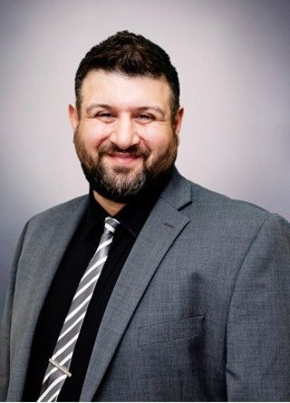 Man in gray suit and tie smiles at the camera. Black shirt, gray and white striped tie, beard.