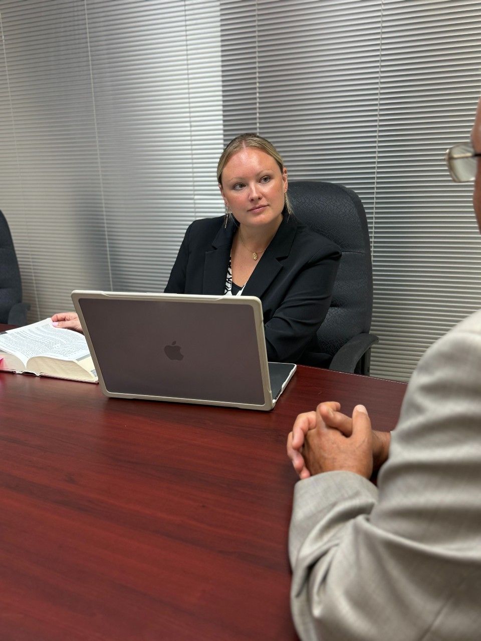 Woman in suit sits at table with laptop, looking at a person out of frame. An open book is to her left.