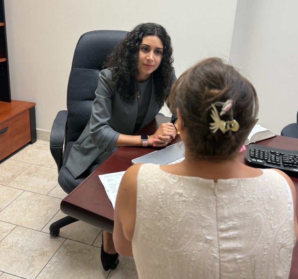 Woman in grey suit sits across desk from another woman. Office setting.
