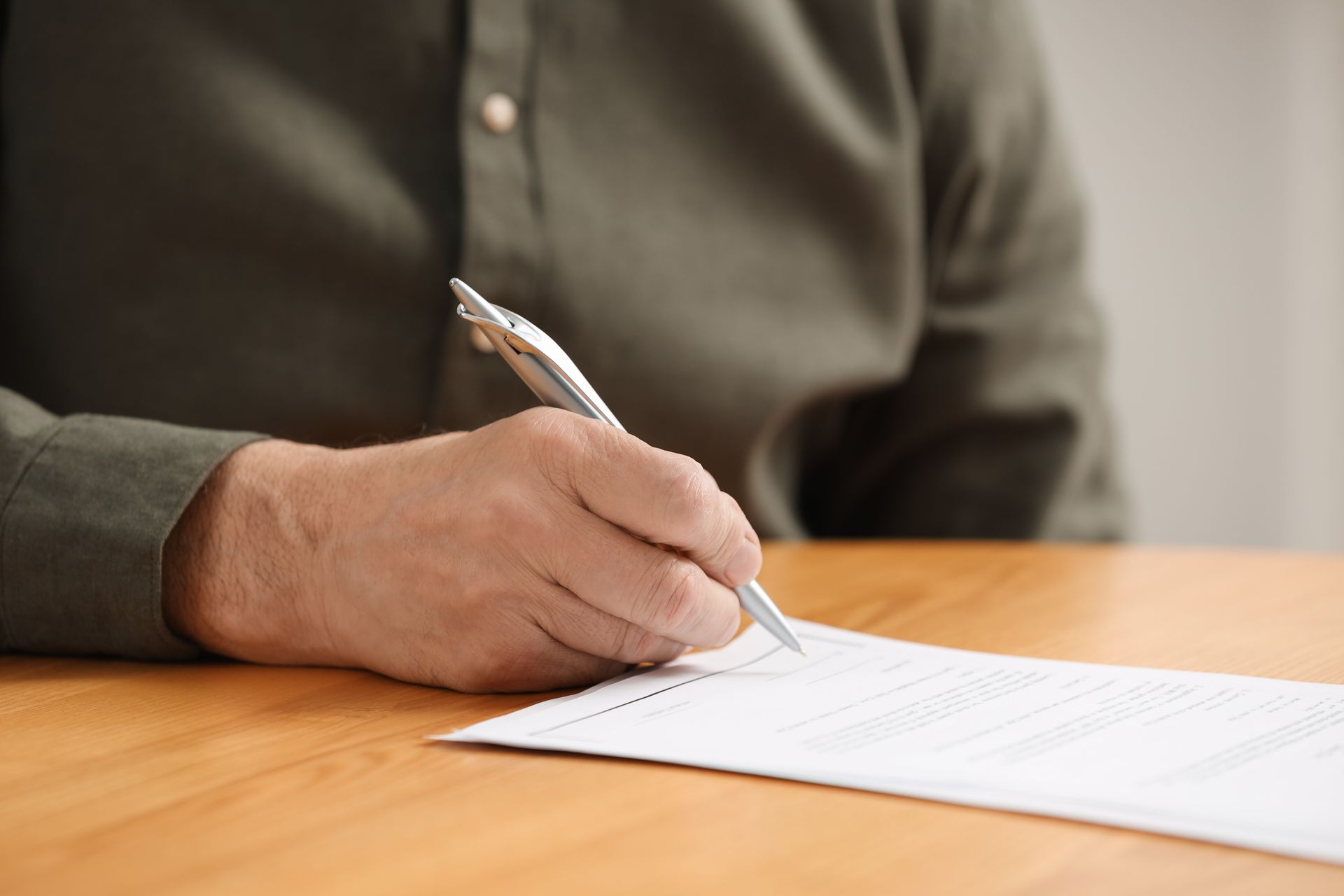Person writing on a document at a wooden table, holding a pen.