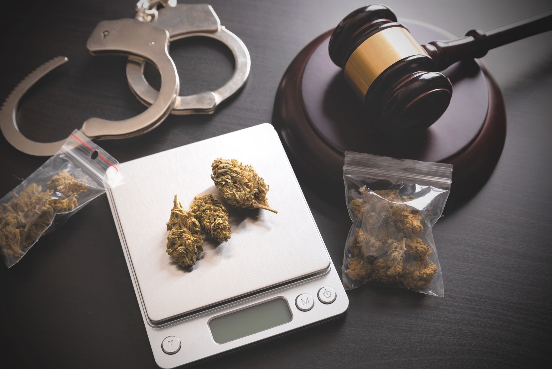 Handcuffs, a judge’s gavel, digital scales, and small bags of cannabis on a dark surface.