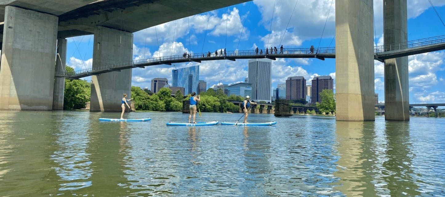 A group of people are standing on paddle boards in the water under a bridge.